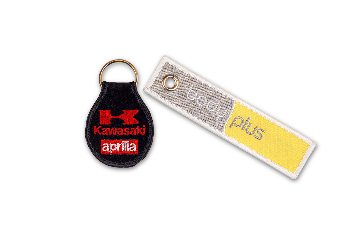Stickabzeichen der Stickerei Tischler als Schlüsselanhänger Schwarzes Stickabzeichen mit roter Aufschrift Kawasaki Aprilia und gelb-graues Stickabzeichen mit der Aufschrift body plus mit Ösen für den Schüsselbund